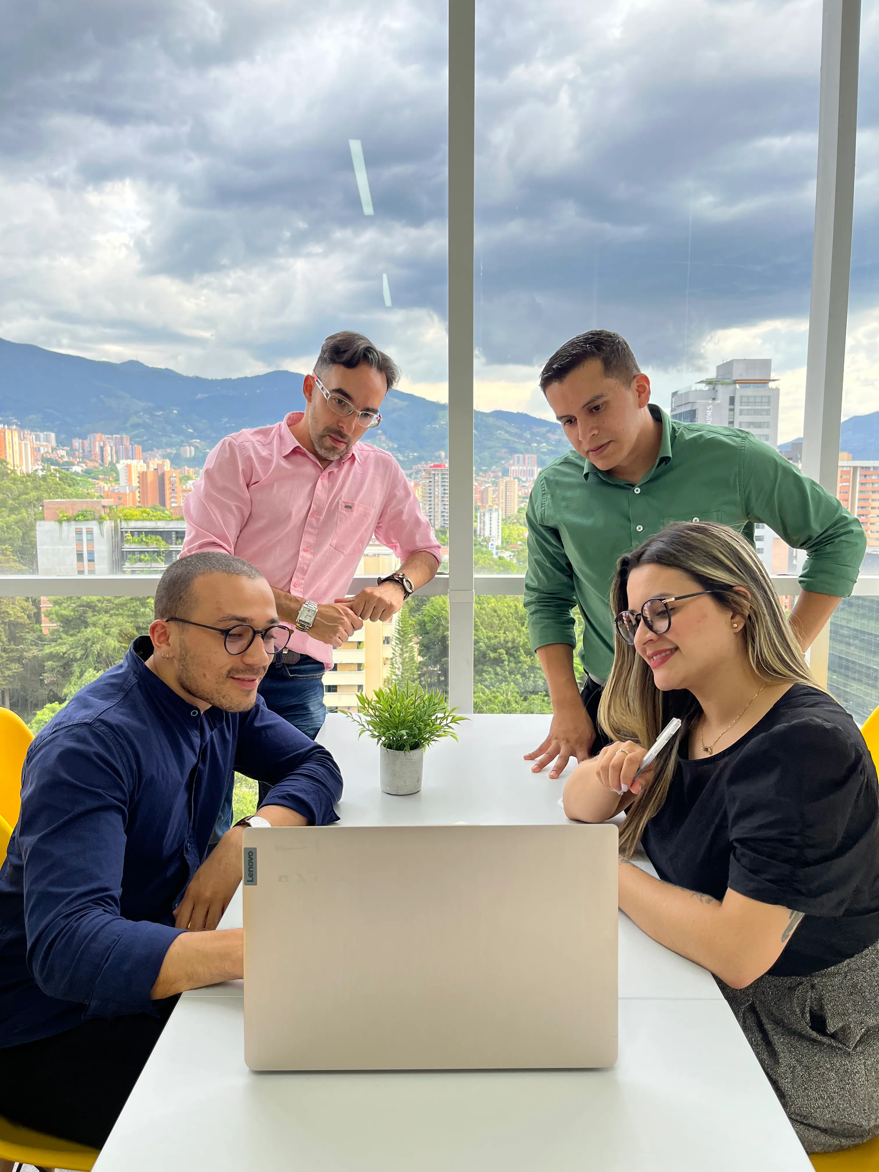 Four Callbox colleagues gathered around a laptop in a bright office with the Medell\u00edn skyline behind them